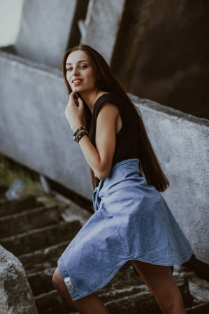 Portrait of a beautiful young brunette woman with long straight hair, wearing a black T-shirt and blue jeans skirt, posing on the stairs.の写真素材