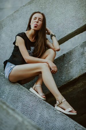 Portrait of a beautiful young woman in jeans shorts and a black T-shirt sitting on the stairs
