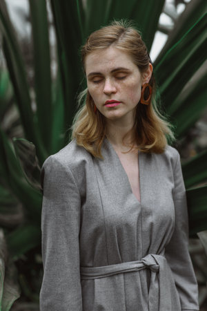 A girl poses in a botanical garden among plants. The model is wearing a gray suit. Freckles on the girl's face. The girl poses among flowers, bushes, trees, cacti.の写真素材