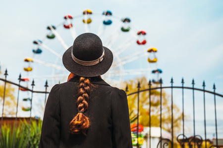 Back view of a young girl in hat standing in front of the ferris wheel at the amusement parkの写真素材