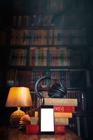 Grouped Mobile phone, headphones and a stack of books near the lamp on a wooden table. Concept of training and audiobooks. Library bookcase in the background with light. Copy space. Mock up.の写真素材