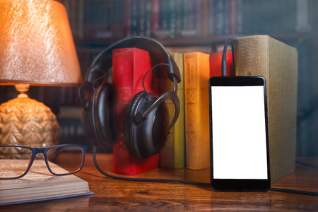 On a wooden table next to the lamp are headphones, a phone, a stack of books and glasses. The concept of modern technology and audiobooks. Mock up.の写真素材
