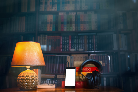 On a wooden table next to the lamp are headphones, a phone, a stack of books and glasses. The concept of modern technology and audiobooks. Tint and copy space.の写真素材