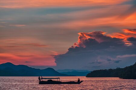 Landscape with a boat floating on the water. On the background of the contour of the shoreline and a fiery sunset. Close up.の写真素材