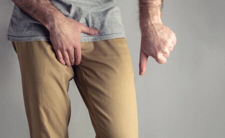 A man in beige jeans holds his genitals and points with his thumb down. Disease for men. The concept of protection against sexually transmitted infections. Testicular cancer.の写真素材
