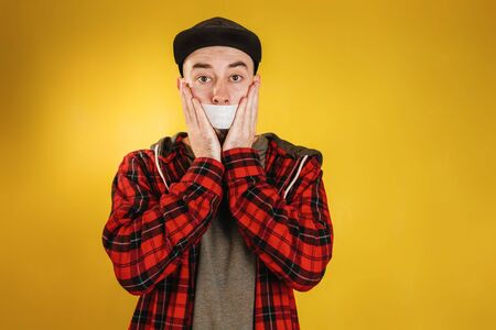 People and emotions. A man in a black cap and a checked red shirt, holding his hands to his face, on which his mouth is taped. Yellow background. Copy spaceの写真素材