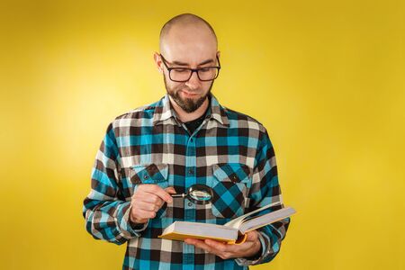 Research and business. A bald man with a beard and glasses, holding a book in his hands and carefully studying it through a magnifying glass. Yellow background. Copy space.の写真素材