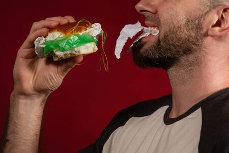The concept of fast food and ecology. A bald man with a beard, happily eating a hamburger stuffed with garbage. Burgundy background. Close up.の写真素材