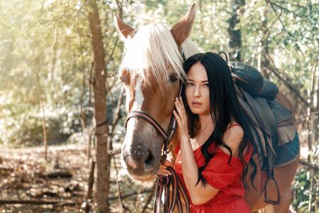 Young beautiful brunette woman in red dress posing near horse.の写真素材