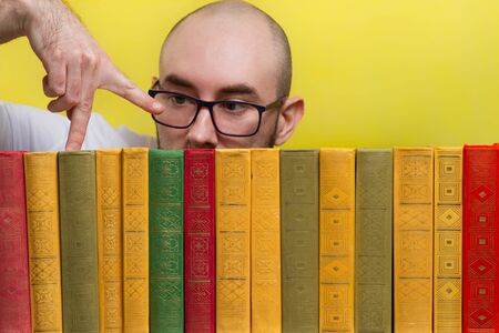 The concept of education and reading. Portrait of a bald man with glasses, who fingers through the books standing in a row. Yellow background. Close upの写真素材