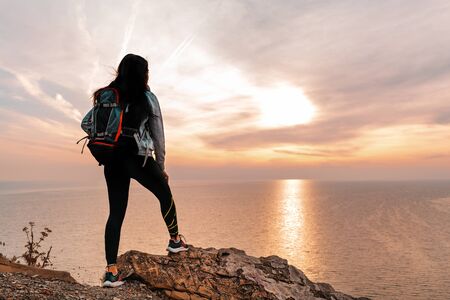 The concept of Hiking and sports recreation. A woman with a backpack on her back poses against the sea and sky. Sunset. Rear view. Copy space.の写真素材