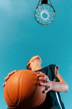 Sports and basketball. A young teenager in a black tracksuit stands with a ball in his hands and prepares to throw the ball into the ring. Blue sky with hoop. Bottom view. Copy spaceの写真素材