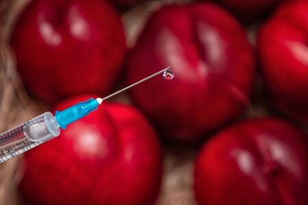 Syringe with chemical injection close-up. Blurred peaches in the background. The concept of GMO foods, vegetables and fruits rainbow.の写真素材