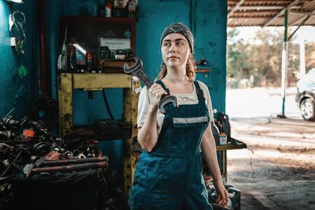 The concept of small business, feminism and womens equality. A young woman in overalls poses with a large wrench and looks awayの写真素材