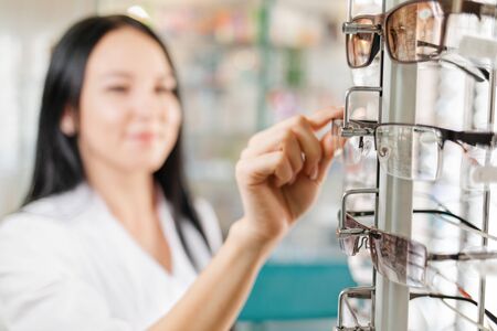 Medicine, pharmacy and optics. A brunette woman in a white coat takes off her glasses with her hands from the window. Close-up of glasses. Blurred backgroundの写真素材