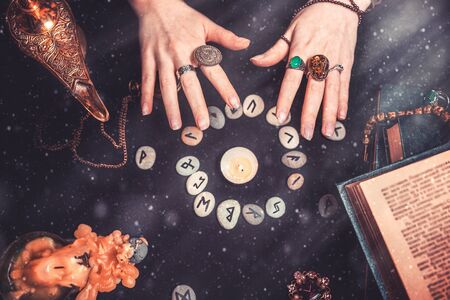 Astrology and esotericism. Female witch hands conjure over the rune circle. On a black background lie fortune-telling runes, a book, precious amulets, a copper lamp and a candle. Dust and light.の写真素材