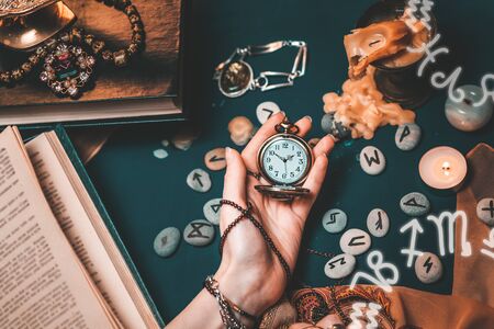 Astrology and esotericism. The female hand of the witch holds a watch on a chain. In the background, old books, fortune-telling runes, candles, and jewels. The view from the top. Zodiac of horoscope.の写真素材
