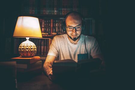 Reading and education. A man in headphones at the library reading a book in the evening by the light...