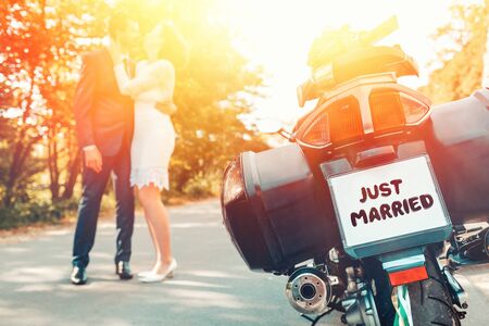 Wedding, newlyweds. The back of a bike with a sign just married. A man and a woman in wedding dresses hug and kiss standing on the road, in a blur in the background. Light and copy space.の写真素材