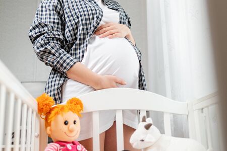 A young pregnant woman stands next to a baby cradle with toys, and hugs her belly. Bottom view....