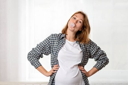 A young pregnant woman shows off her tummy and inflates a bubble of gum. White window in the...