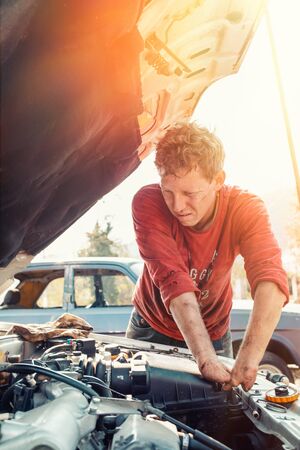 The concept of disability of people and their adaptation to life. Blond disabled man repairs the engine of the car. There are no fingers on his hands. Vertical. Light.の写真素材