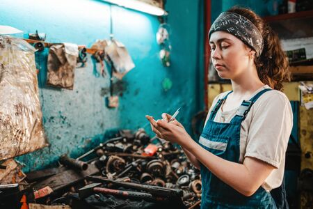 Gender equality. Portrait of a young woman in uniform, working in a workshop, which writes down orders on a piece of paper. In the background is a blue wall and a table with spare parts for the car.の写真素材