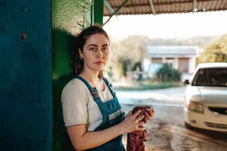 Gender equality. A portrait of a young brunette in uniform leaning against the gate and wiping her hands with a rag. In the background, a street and a white car.の写真素材