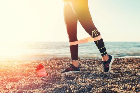 The concept of sport. A young woman exercises with a power band on her legs. There's a pink shaker on the ground. In the background, the sea and the horizon line. Close up. Copy space and sunlight.の写真素材