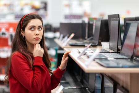 A young brunette woman in a red sweater thoughtfully selects a laptop in the store. Close up. The...