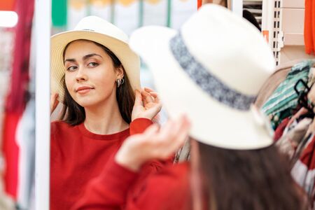 A young Caucasian woman tries on a straw hat for a vacation, and looks in the mirror. The concept of buying clothes.の写真素材