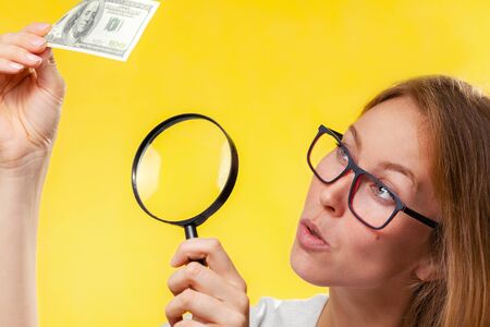 The concept of checking counterfeit money. A blond woman with interest in looking at a dollar bill through a magnifying glass, checking for falsity. Yellow background.の写真素材