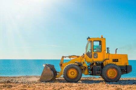 Yellow bulldozer on the background of clear blue sky and seashore. Copy space and light from the...