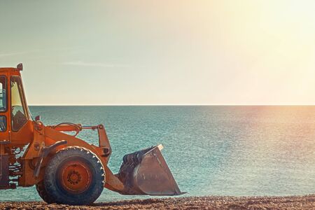 Yellow bulldozer rides on the beach. In the background sea and sky. Light from the right side.
