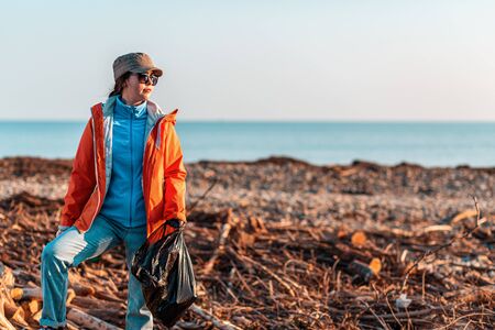 A young Caucasian female volunteer in a jacket, posing with a plastic garbage bag in her hands, posing against a dirty beach, sea and sky. Concept of environmental pollution. Copy space.の写真素材