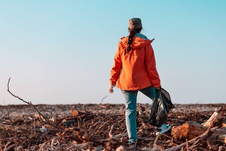 A young Caucasian female volunteer in a jacket stands with a garbage bag in her hands. In the background, a muddy beach and the sky. Concept of environmental pollution. Copy space.の写真素材