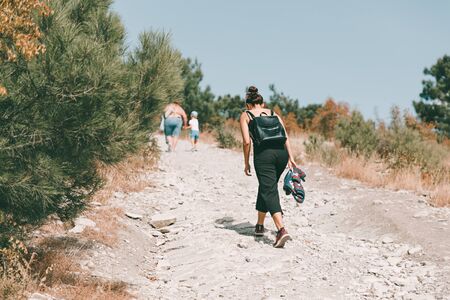 Tourism and outdoor activities. A woman with a backpack on her back is walking along a dusty country road. In the background, people walking forward to the forest.の写真素材