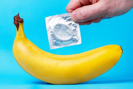 Yellow banana and a man's hand holding a package with a condom, on a blue background. The concept of safe sex.の写真素材