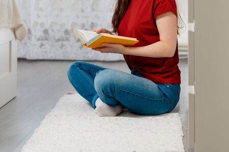 A woman sits cross-legged on the floor and holds a book in her hands. Close up. In the background, the white interior of the room. The concept of education and reading books.の写真素材
