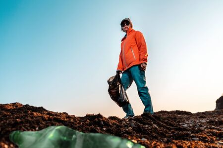 A young Caucasian female volunteer in a jacket, posing with a large garbage bag. Bottom view. The...