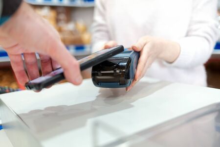A woman's hands are held by a payment terminal and a man pays for a purchase using a smartphone. Copy space. The concept of NFC, business and banking transactions.の写真素材
