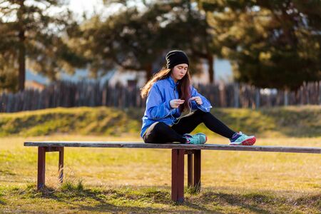 A young Caucasian woman in sportswear sits on a bench and untangles the headphones from the player....