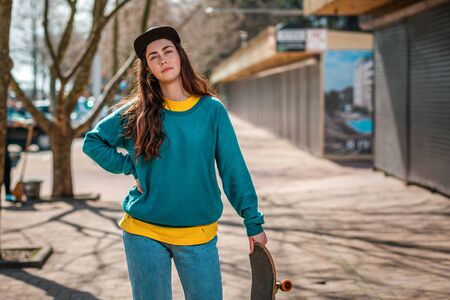A young Caucasian hipster woman poses with a skateboard. In the background, an alley. Close up....