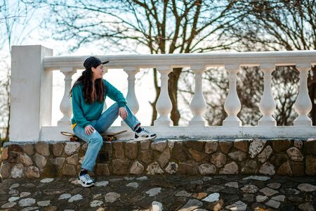 A beautiful hipster teenage girl sitting on a skateboard and looking into the distance. In the...
