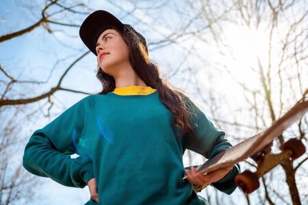 A young caucasian hipster woman poses with a skateboard in her hands and looks into the distance. In the background, blue sky, trees and sun. Bottom view. Concept of sports lifestyle and street culture.の写真素材