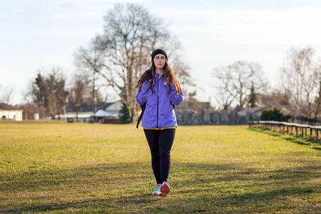 A young Caucasian woman in sportswear with a backpack behind her is walking on the lawn.