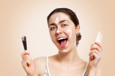 Portrait of a young happy woman with a smile using a mask for skin care. Beige background. Concept of cosmetology, cosmetics and skin care.の写真素材