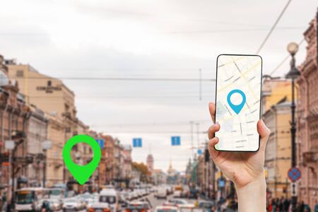 A woman's hand holds a smartphone with an online map app. In the background is a city street with traffic, with a green location icon.Concept of online navigation and GPS.の写真素材