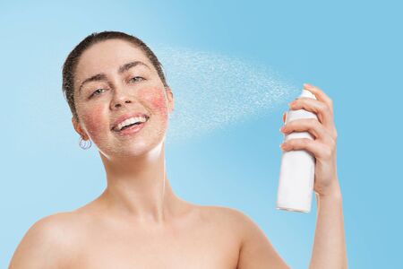A young Caucasian woman with rosacea on her cheeks splashes thermal water on her face and smiles with relief. Blue background. Concept of cosmetology, skin care and rosacea.の写真素材