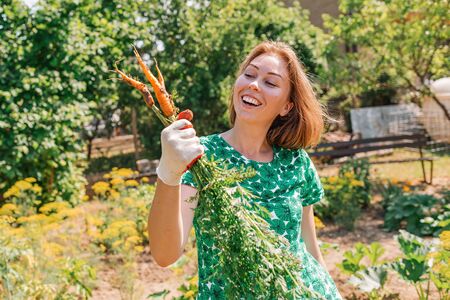 A Caucasian young woman joyfully holds a bunch of carrots collected from the garden.Vegetation in the background.Concept of harvesting and gardening.の写真素材